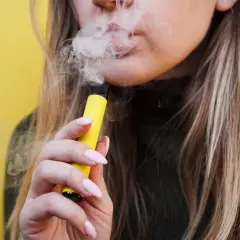Mujer vapeando un dispositivo amarillo, liberando humo. El vaper pone los dientes amarillos, afectando la sonrisa.