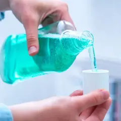 Mano vierte líquido azul de una botella a una taza medidora, mostrando varios tipos de clorhexidina para higiene bucal.