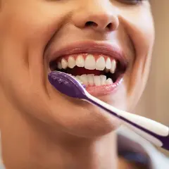 Primer plano de una mujer sonriendo mientras se lava los dientes con un cepillo morado.