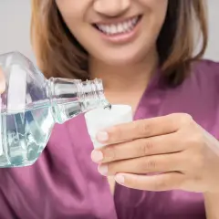 Mujer sonriente vierte líquido de una botella a una taza. Podría ser clorhexidina o agua con sal para enjuague bucal.