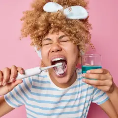 Mujer con rizos se cepilla los dientes y sostiene un vaso con enjuagues para periodontitis.