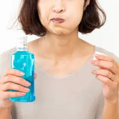 Mujer con mejillas llenas de líquido azul, sosteniendo una botella de enjuague bucal con antibiótico y un tapón.