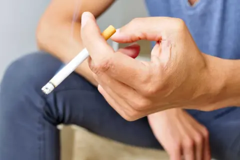 Mano sujetando un cigarrillo encendido. Fumar aumenta el riesgo de rechazo de implantes dentales.