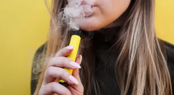 Mujer vapeando un dispositivo amarillo, liberando humo. El vaper pone los dientes amarillos, afectando la sonrisa.