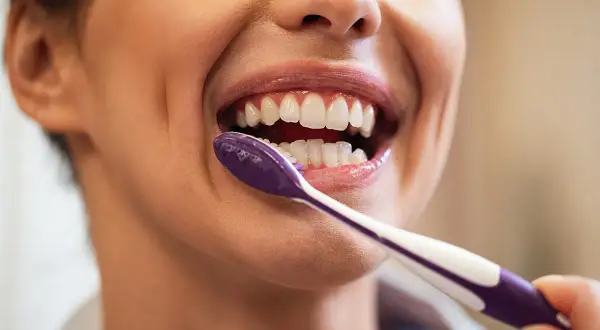 Primer plano de una mujer sonriendo mientras se lava los dientes con un cepillo morado.