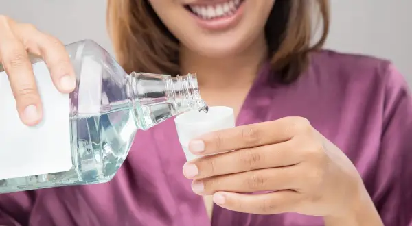 Mujer sonriente vierte líquido de una botella a una taza. Podría ser clorhexidina o agua con sal para enjuague bucal.