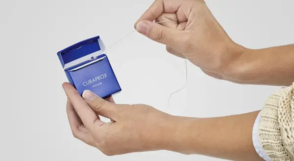 Manos sacando hilo dental de caja azul Curaprox. ¿El hilo dental se pasa antes o después del cepillado?