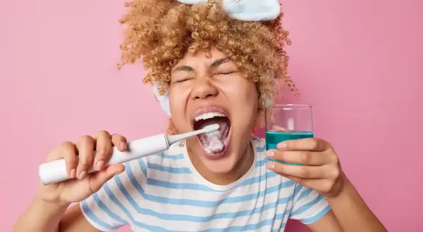 Mujer con rizos se cepilla los dientes y sostiene un vaso con enjuagues para periodontitis.