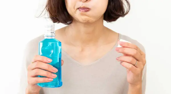 Mujer con mejillas llenas de líquido azul, sosteniendo una botella de enjuague bucal con antibiótico y un tapón.