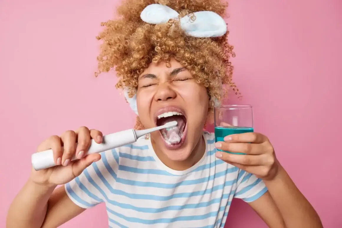 Mujer con rizos se cepilla los dientes y sostiene un vaso con enjuagues para periodontitis.