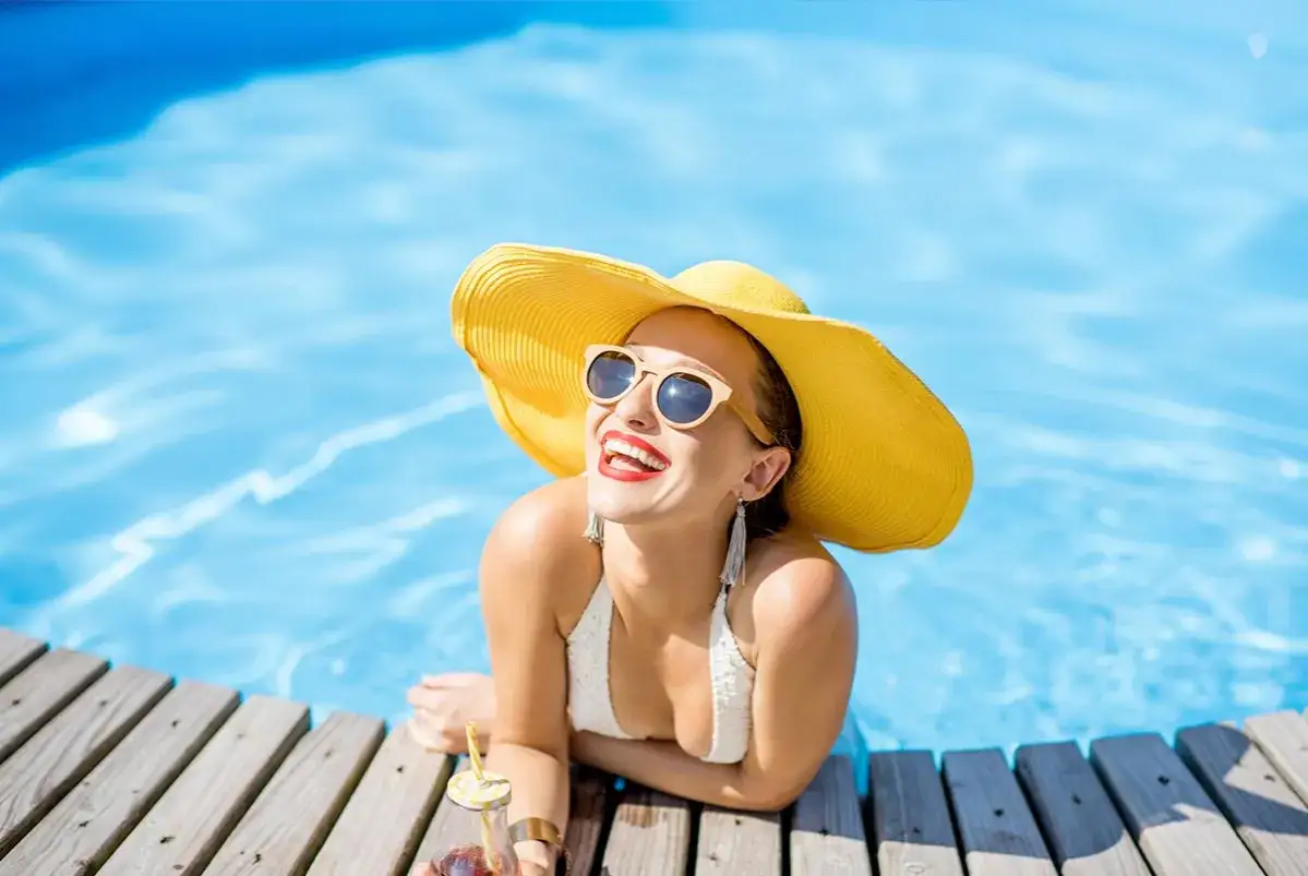 Mujer sonriente con sombrero amarillo y gafas de sol, disfrutando de la piscina. &iexcl;Un verano para proteger tus dientes con una sonrisa radiante!