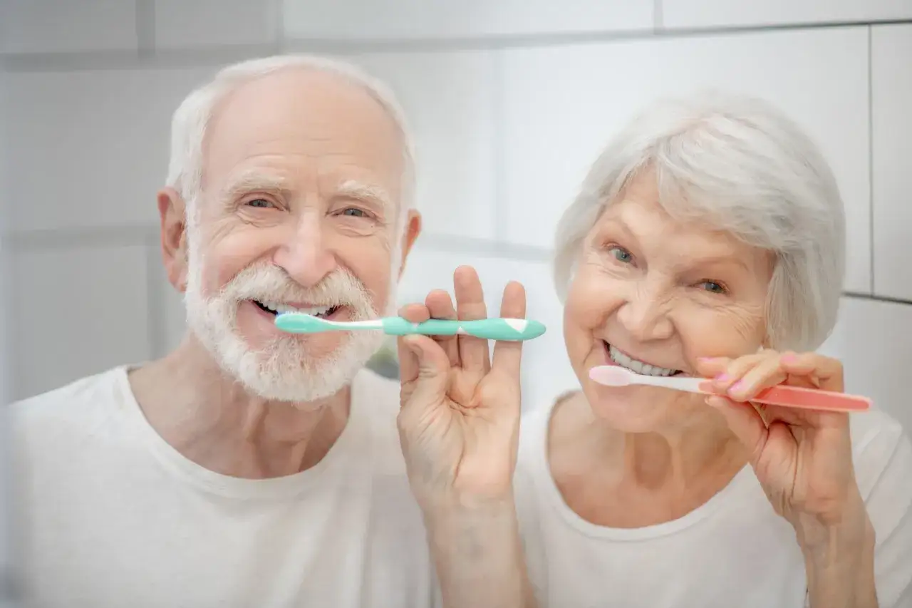 Pareja mayor sonriendo y cepillándose los dientes juntos, demostrando la importancia del cuidado de los dientes a cualquier edad.