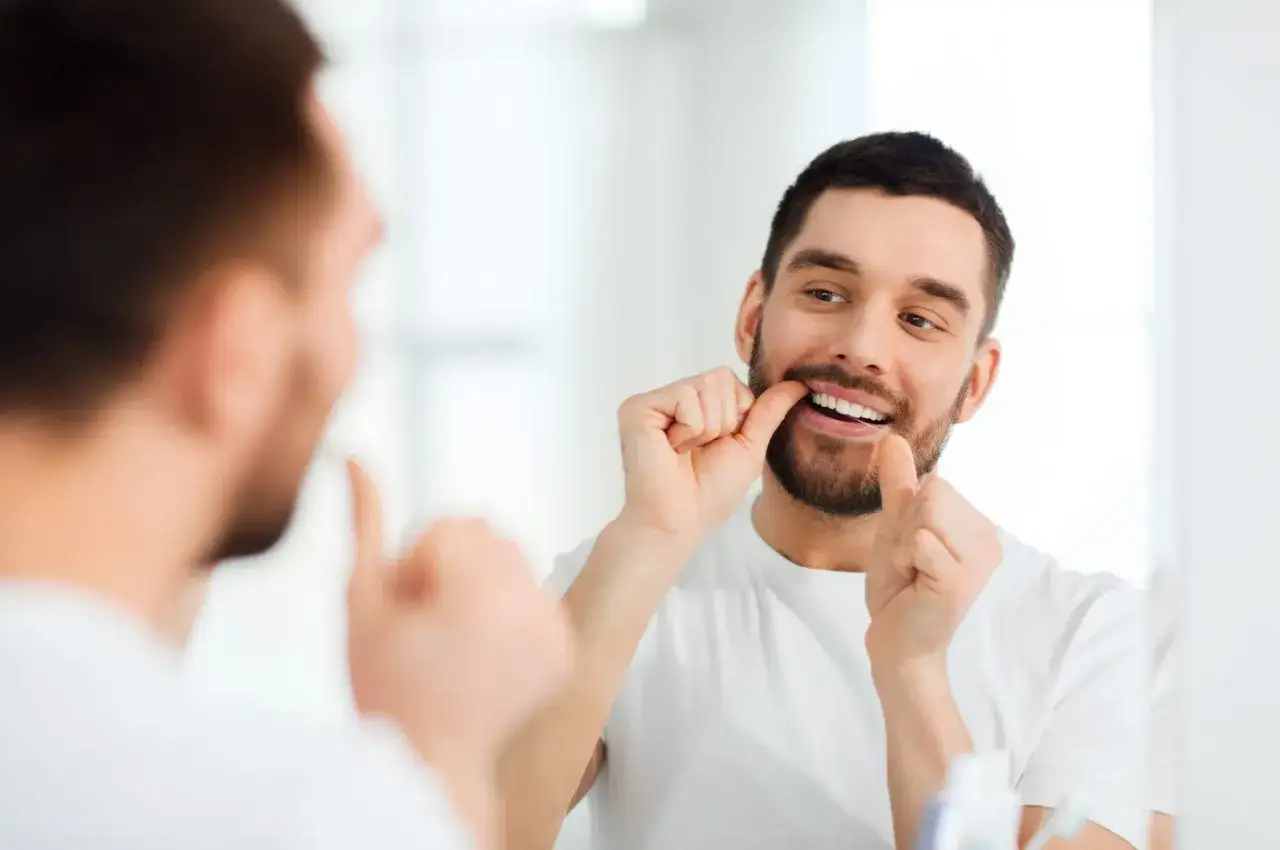 Hombre sonriente se pasa el hilo dental frente al espejo. La pregunta es si el hilo dental se pasa antes o despu&eacute;s del cepillado.