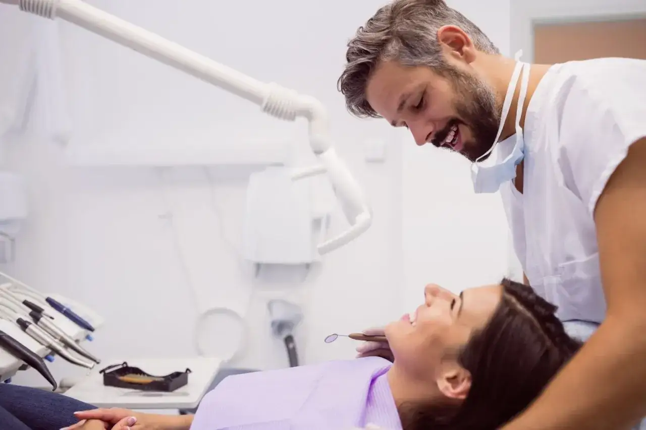 Dentista sonriente revisa la boca de una paciente, indicando una consulta para fluorizaci&oacute;n dental en adultos.