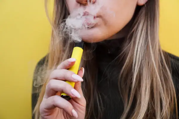 Mujer vapeando un dispositivo amarillo, liberando humo. El vaper pone los dientes amarillos, afectando la sonrisa.
