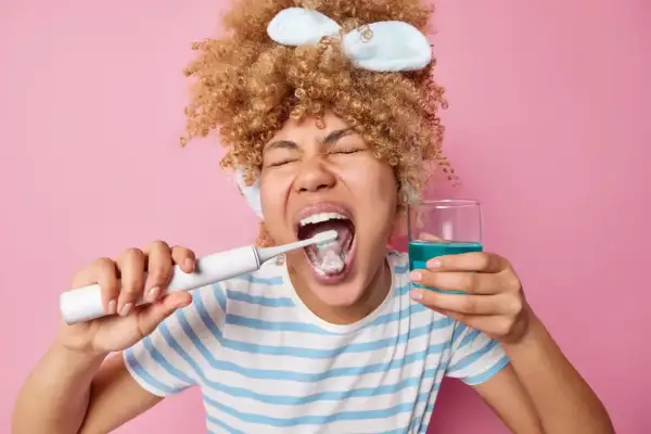 Mujer con rizos se cepilla los dientes y sostiene un vaso con enjuagues para periodontitis.