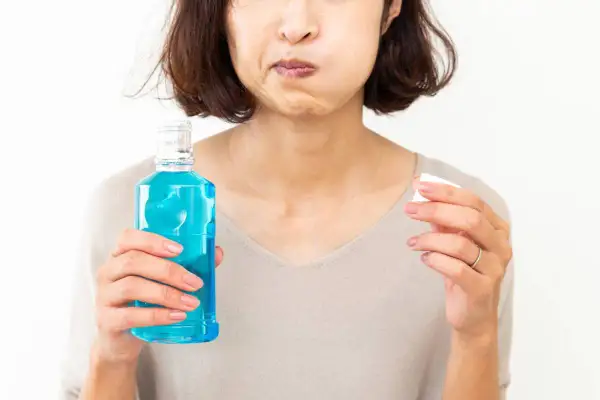 Mujer con mejillas llenas de líquido azul, sosteniendo una botella de enjuague bucal con antibiótico y un tapón.